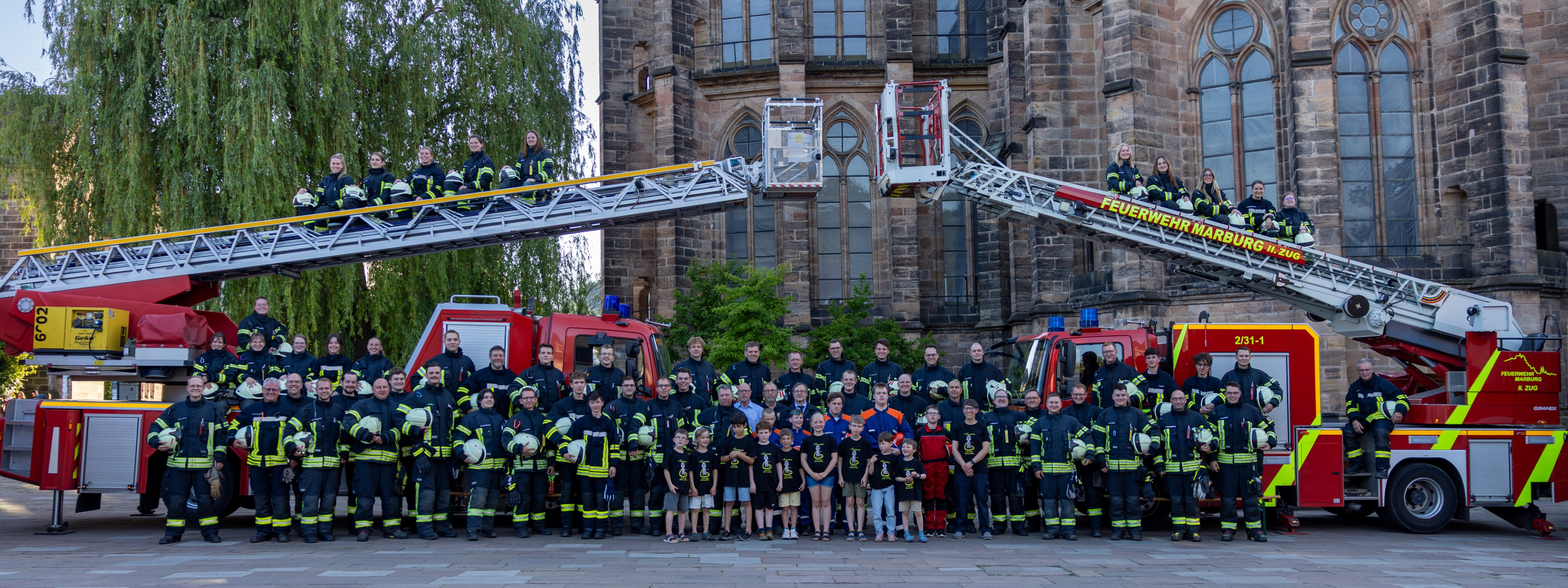 Großes Gruppenfoto der Feuerwehrleute in Einsatzkleidung vor einem historischen Gebäude mit zwei ausgefahrenen Drehleitern
